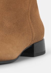 Bottine en daim marron à finition texturée, silhouette arrondie, et talon carré noir bas. Présente des coutures lisses sur les côtés.