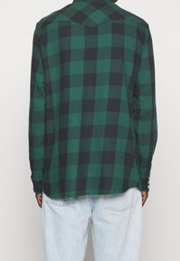 Zielona i czarna koszula w kratę z długim rękawem, z wydłużonym kołnierzykiem i guzikami na mankietach, pokazana z tyłu.