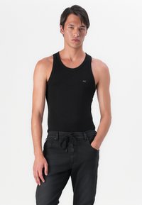 Črn brezrokavnik iz mehkega in raztegljivega materiala. Ima okroglo ovratnico in prilegajočo se zasnovo. Oblečen z temnimi kavbojkami.