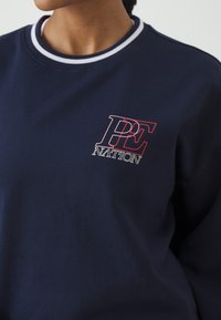 Sweat-shirt bleu marine avec logo "PE Nation" blanc et rose sur la poitrine et col côtelé à rayures blanches porté par une personne.
