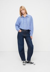 Φωτεινό γαλάζιο ριγέ πουκάμισο με βολάν στο εμπρόσθιο μέρος, συνδυασμένο με ψηλόμεσα σκούρα μπλε τζιν και μαύρα παπούτσια Converse.