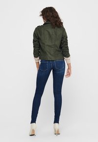Veste en suédine verte olive avec un design ajusté, des détails aux épaules et des poignets côtelés, associée à un jean slim en denim brut.