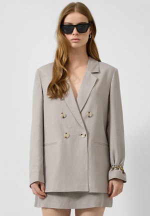 Femme aux longs cheveux bruns portant des lunettes de soleil noires en œil de chat et un blazer gris clair oversize à double boutonnage avec une jupe.