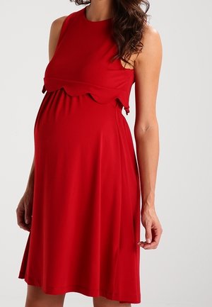 Kobieta w ciąży w czerwonej, bezrękawnikowej sukience ciążowej z falowanym detalem pod biustem na jednolitym tle.