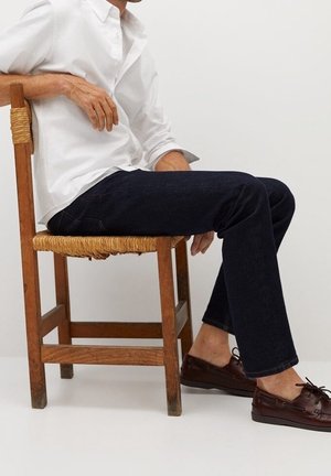 Jean en denim bleu foncé à coupe droite, associé à une chemise blanche boutonnée et des chaussures bateau en cuir marron, assis sur une chaise en bois.