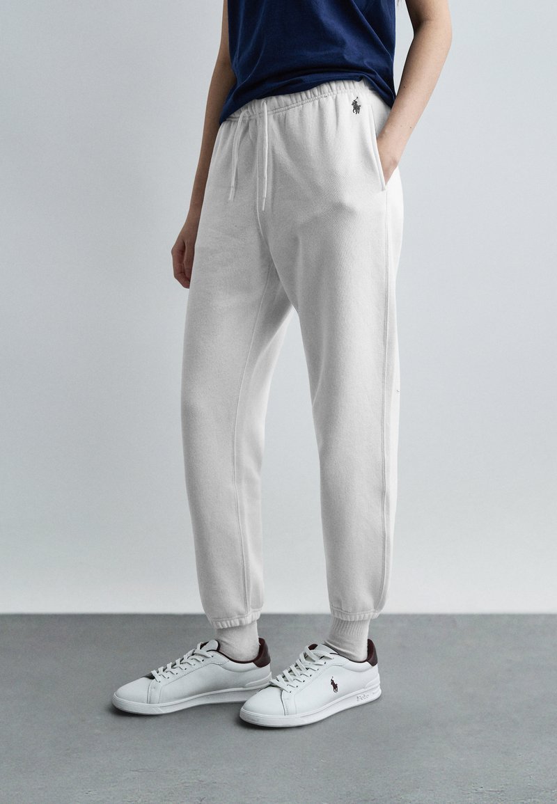 Άτομο που φοράει λευκή φόρμα με ελαστικά μανσέτα και λευκά αθλητικά παπούτσια με σκούρες λεπτομέρειες στη φτέρνα, στέκεται σε γκρι δάπεδο με απλό φόντο.