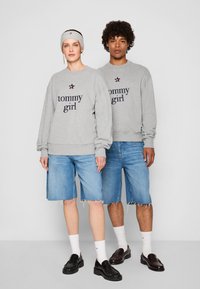 Felpe grigie con "tommy girl" in blu navy, abbinate a pantaloni corti di jeans. Scarpe nere e calze bianche completano gli outfit. Design semplice con un look casual.