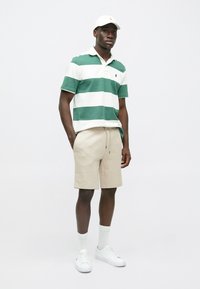 Zelená a biela pruhovaná polokošeľa, béžové šortky, biele tenisky a ponožky. Voľný strih a mäkký materiál. Jednoduchý, čistý dizajn.