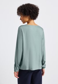 Blusa a maniche lunghe in morbido tessuto teal chiaro con vestibilità rilassata, scollo a barca e polsini elastici. Caratterizzata da una texture liscia e un design minimale.