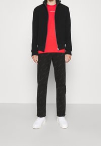 Giacca nera con zip, t-shirt rossa con logo, pantaloni neri a fantasia e scarpe da ginnastica bianche. I materiali includono cotone e miscele sintetiche.