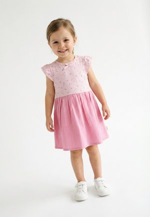 Lächendes junges Mädchen steht, trägt ein rosa Kleid mit floralem Oberteil und weißen Turnschuhen vor einem schlichten weißen Hintergrund.
