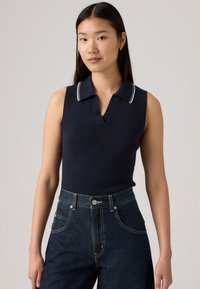 Polo sans manches bleu marine avec texture côtelée et rayures blanches sur le col, associé à un jean en denim sombre taille haute.