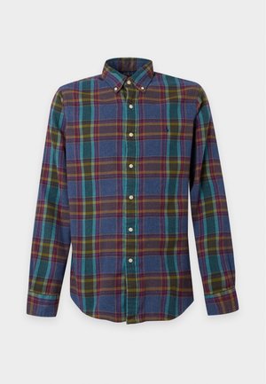 Jeune homme portant une chemise à carreaux bleue et verte à boutons, un pantalon marron foncé et des mocassins beige clair, debout devant un fond uni.