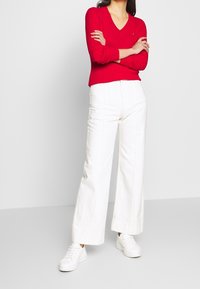 Pull rouge en maille côtelée avec un col en V, associé à un pantalon blanc à jambes larges et des baskets blanches, présentant une tenue décontractée.