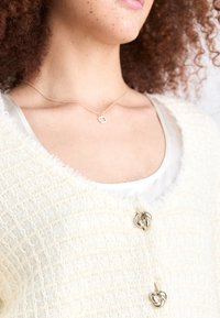 Hvid tekstureret cardigan med frynser, prydet med knapper i sølvfarve. Under er en lys satin top. En simpel halskæde er synlig.