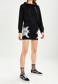 Černá mikina s šňůrkami, džínsová sukně s bílou květinovou výšivkou, doplněná punčocháčemi s rybářskou sítí a černými teniskami s bílými akcenty.