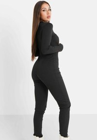 Schwarzer langärmeliger Jumpsuit mit tailliertem Design, gerippter Textur und knöchellangen Beinen. Verfügt über einen hohen Ausschnitt und minimalistischen Stil.