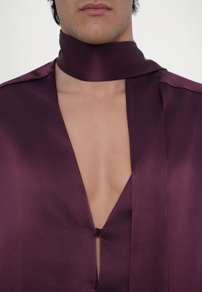 Short purple satin garment with a deep V-neck, featuring two buttons at the chest and a matching fabric neck tie. Smooth texture, minimal design.