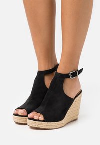 Černé semišové sandály na klínu s otevřenou špičkou, popruhem na kotníku a tkanou jutu platformou. Mají hladkou texturu a elegantní design.