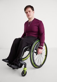 Fekete vázú, zöld akcentusokkal ellátott manőverezhető kerekesszék. Egyén bordó hosszú ujjú pólóban, fekete nadrágban és fekete cipőben. Egyszerű háttér.