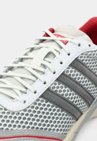 Chaussure de sport grise avec tige en mesh, accents blancs et gris, lacets texturés, détails rouges sur la languette, et trois bandes grises sur le côté.