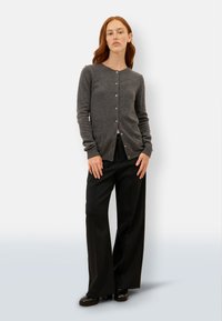 Szary kardigan z długim rękawem, zapinany na guziki, wykonany z miękkiej mieszanki wełny. Posiada okrągły dekolt i dwie kieszenie z przodu. Noszony na czarnych szerokich spodniach.