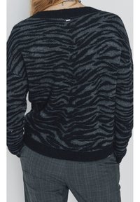 Pull en noir et gris à rayures avec un motif zèbre texturé, col et poignets côtelés. Porté avec un pantalon à carreaux gris.