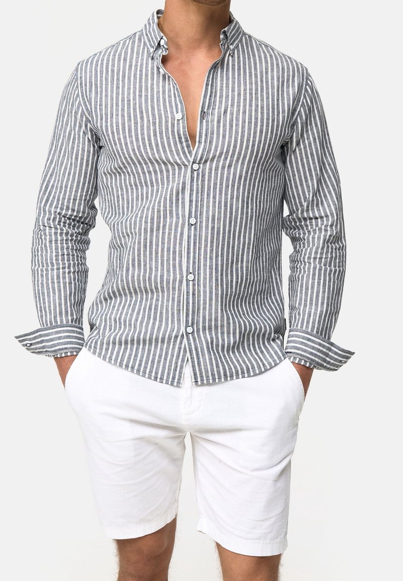 Man draagt een grijs-wit verticaal gestreept shirt met lange mouwen, witte knoopsluiting en witte korte broek, met handen in de zakken, tegen een effen achtergrond.