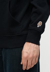 Χέρι που φοράει μαύρο φούτερ με ένα μικρό κεντημένο παγωτό κοντά στον καρπό, συνδυασμένο με σκούρο τζιν παντελόνι.