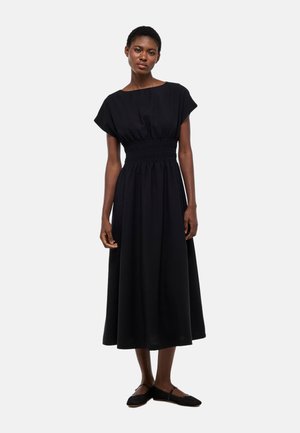 Femme debout portant une robe midi noire à taille froncée et manches courtes, associée à des chaussures plates Mary Jane noires sur un fond blanc.