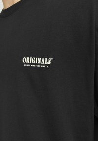 Juodas medvilnės marškinėliai trumpomis rankovėmis, su balta spauda kairėje krūtinės pusėje "ORIGINALS" ir "©NUO DEVYNIASDEŠIMT".