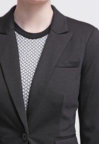 Černý blazer s přiléhavým střihem, na jedno knoflíkové zapínání a s malou náprsní kapsou, vrstvený přes vzorovaný šedý top.