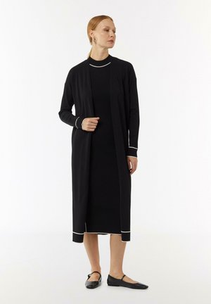 Zwarte lange cardigan over een zwarte ribbed jurk, met witte afwerking bij de mouwen en de zoom. Model staat op zwarte platte schoenen tegen een neutrale achtergrond.