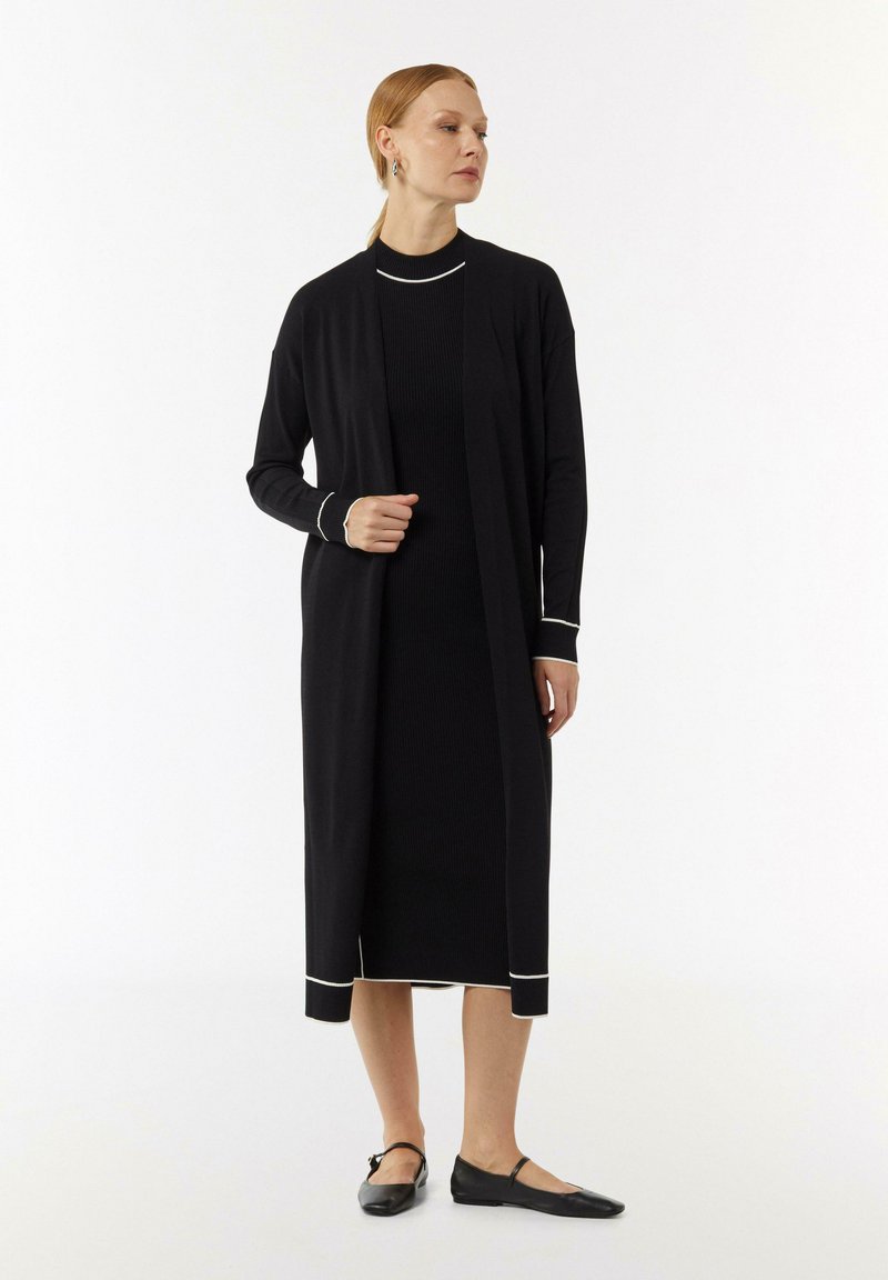 Schwarzer langer Cardigan über einem schwarzen, gerippten Kleid, mit weißem Saum an den Ärmeln und dem Saum. Das Modell steht in schwarzen flachen Schuhen vor einem neutralen Hintergrund.