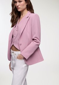 Blazer rosa chiaro con vestibilità su misura, rever a notched e due bottoni. Indossato sopra un gilet rosa corto e abbinato a pantaloni bianchi a vita alta.