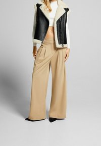 Svart läderväst utan ärmar med crèmefärgad syntetpäls, tillsammans med beiga vida byxor med veck och svarta spetsiga klackar.