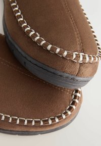 Bruna mockasiner i mocka med vit söm längs med sömmarna och en mörk gummisula. Ovandelarna är mjuka och strukturerade.