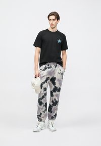 Svart t-shirt med korta ärmar och en blå stjärnlogotyp, kombinerad med tie-dye-byxor i grå och svarta toner. Vita sneakers och en beige keps hålls i handen.