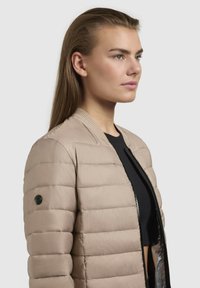 Giacca imbottita beige con colletto a costine e design aderente. Presenta una zip nera e un dettaglio con il logo sulla manica sinistra. Tessuto liscio.