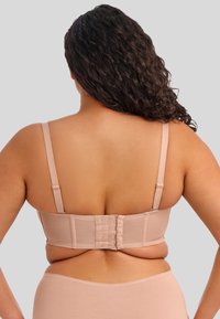 Sujetador beige con tirantes ajustables, cierre de gancho y ojo, y una textura suave. Banda visible y tejido color piel sobre un fondo gris.