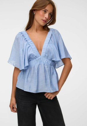 Femme portant un chemisier bleu clair à col en V avec manches volantées et taille, assorti à un pantalon noir, sur un fond uni.