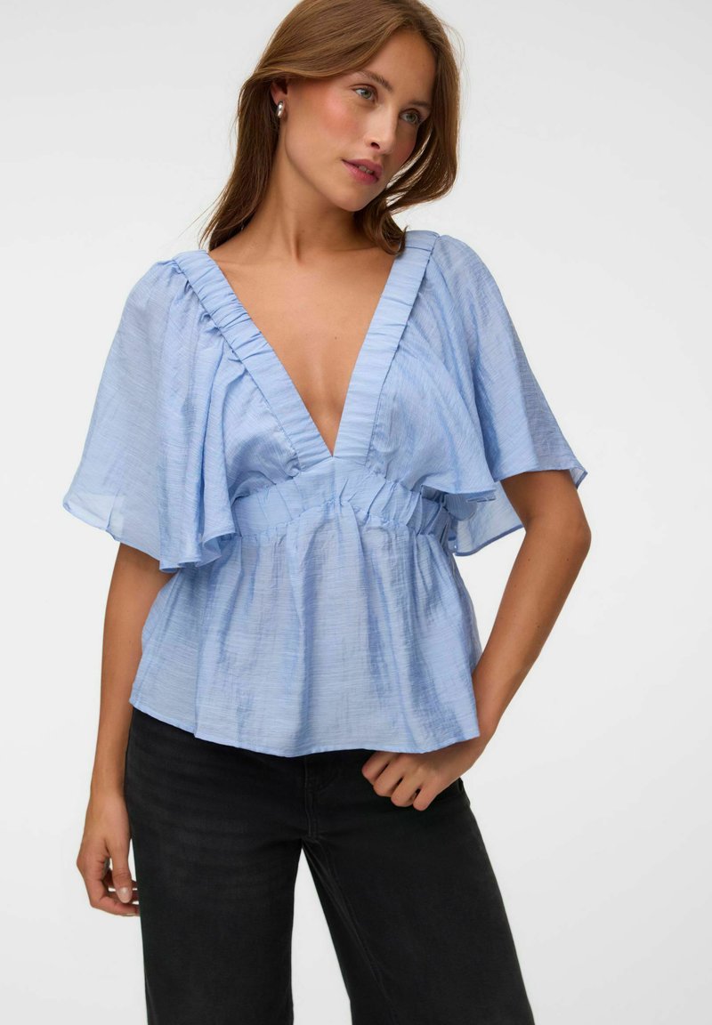 Femme portant un chemisier bleu clair à col en V avec manches volantées et taille, assorti à un pantalon noir, sur un fond uni.