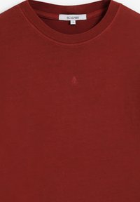 T-shirt rouge rouille à col rond avec un petit logo brodé au centre et une étiquette de marque Scalpers à l'intérieur du col.