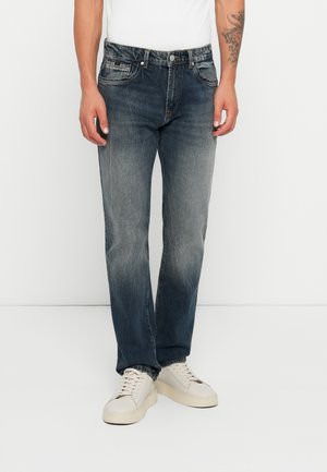 Jeans azules descoloridos con corte recto, cinco bolsillos y costuras en contraste, combinados con zapatillas blancas. Textura de denim suave.