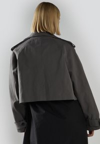 Lyhyt harmaa trench-takki, jossa on rakenteellinen muotoilu, suuri kaulus ja napilliset hihansuut, valmistettu teksturoidusta kankaasta.