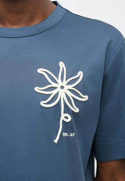 T-shirt de algodão azul com um design de flor bordado em branco do lado esquerdo e o texto "NN. 07" abaixo da flor. Mangas curtas.