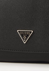 Borsa in pelle nera con una texture, che presenta un logo triangolare in metallo oro e nero di Guess con un accento a punto interrogativo.