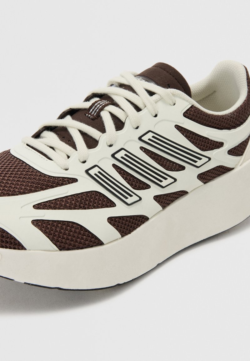 Braune Mesh-Sneaker mit weißen Synthetik-Akzenten, ausgestattet mit einer dicken Sohle und schwarzen Details, die ein modernes, sportliches Design zeigen.