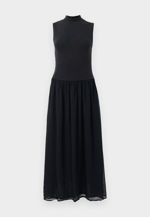 Ilmaõlga must maksikka, millel on vale kaelus, keha külge liibuv ülaosa ja kokku pandud seelik, lihtsa valge tausta ees.