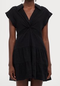 Svart klänning med korta luvaärmar, krage och knuten detalj fram. Har tierade volanger vid kjolen för ökad volym.